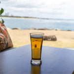 The Polycarbonate Pilsner Glass is full of beer and is sitting on a table in front of the beach. This glass is a great addition to the booze towers beer tower